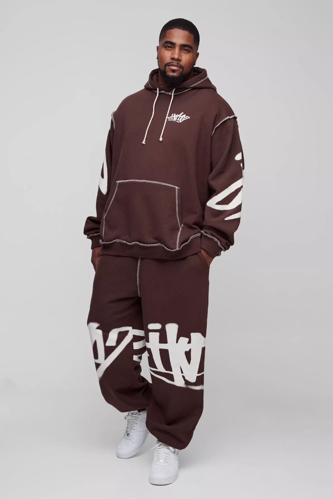 streetwear dark brown tracksuit — hooded, drop shoulder, exposed white stitching, white rope drawstrings, large white graffiti-style graphic print, kangaroo pocket