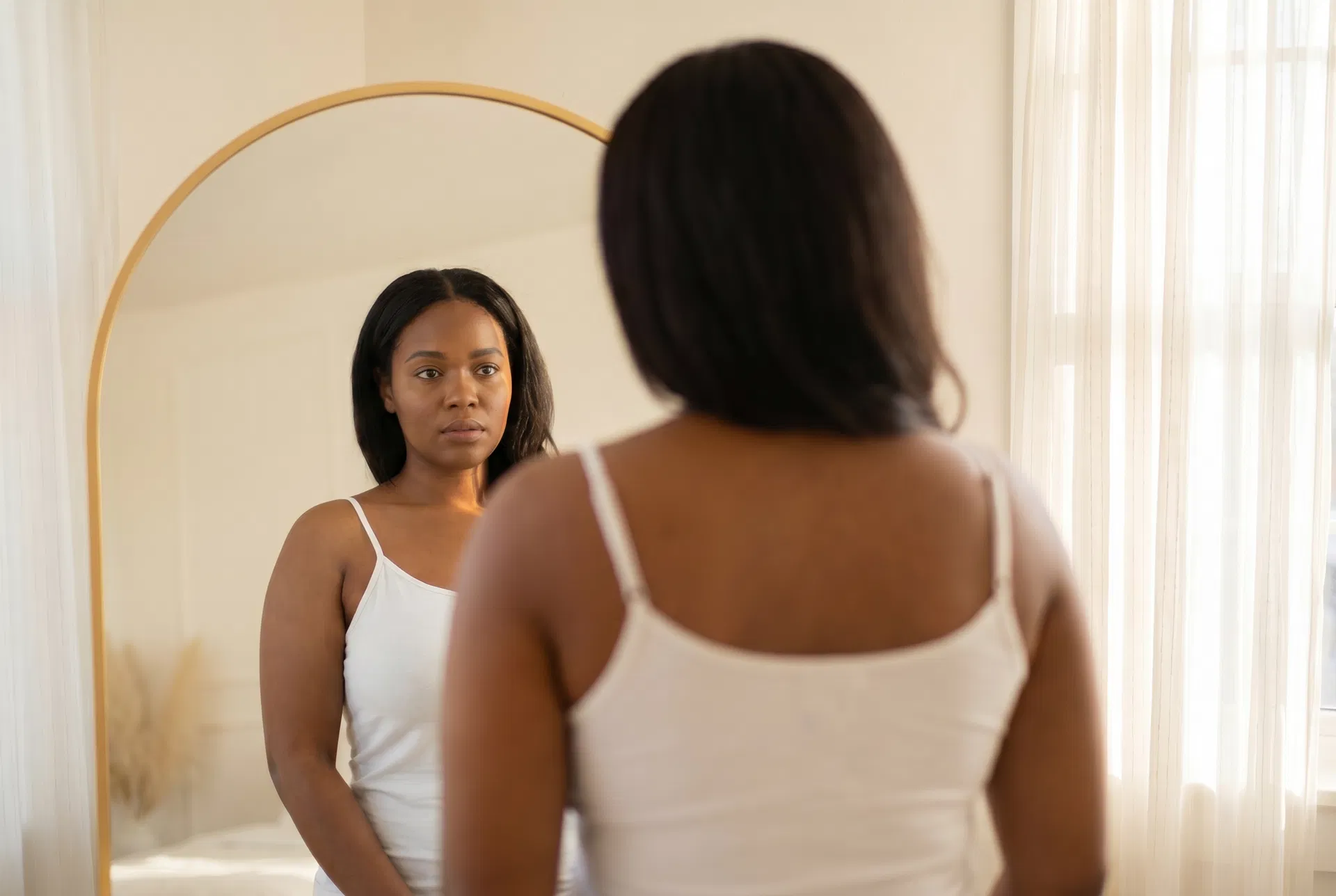 A woman of color looking at her reflection in a gold arched mirror with a neutral, contemplative expression