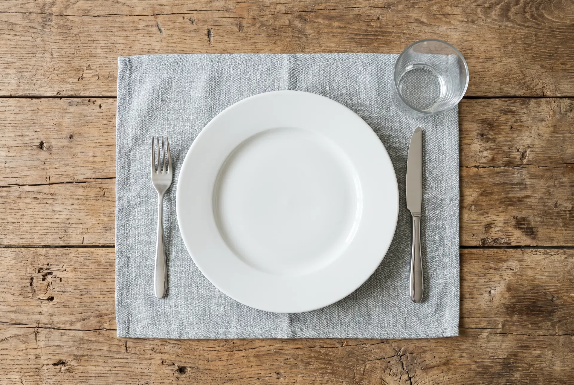 Minimalist place setting with empty white plate, fork, knife, and drinking glass on pale grey-blue linen placemat on weathered wood table