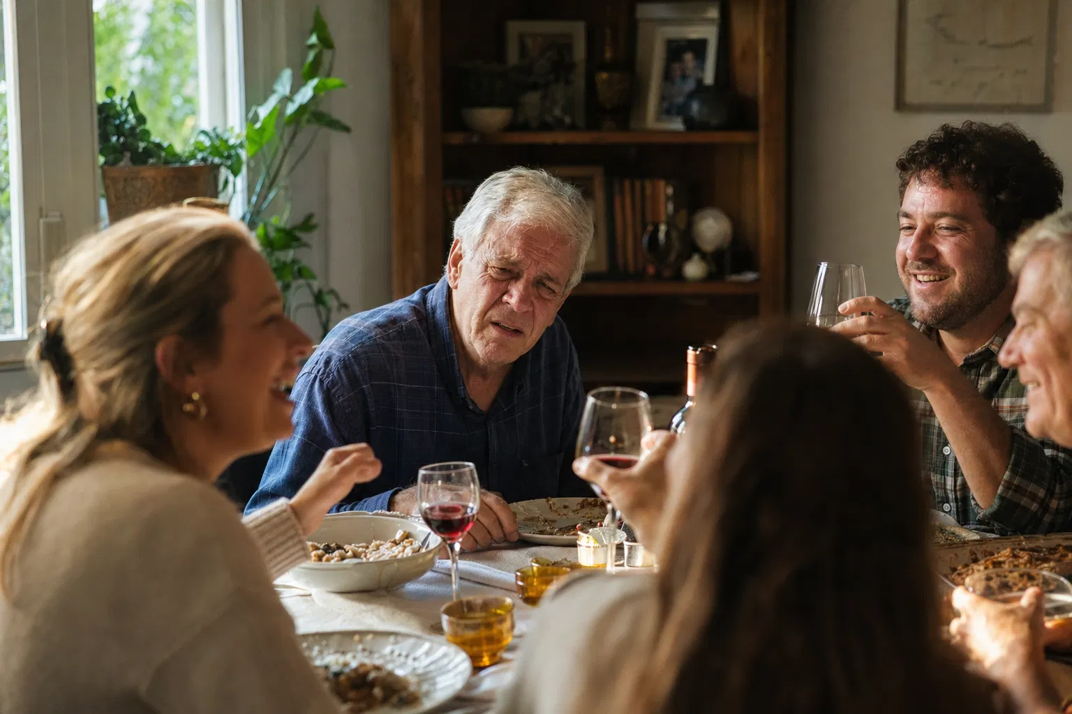Senior man at a family dinner table, leaning forward with a confused, mentally strained expression as relatives laugh around him.