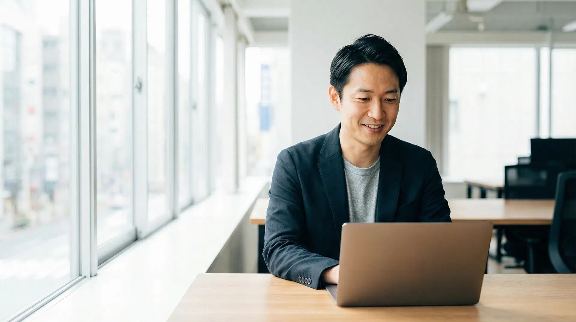 A professional Asian man working at a laptop in a bright modern office, no glasses, relaxed and focused