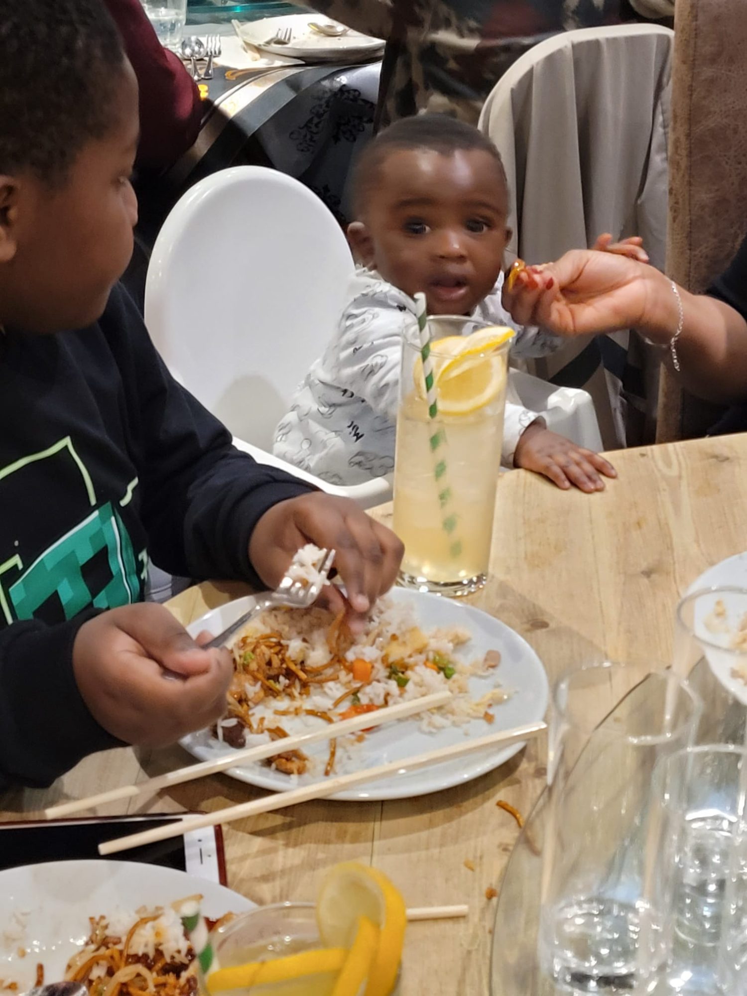 Boy eating rice beside a toddlerin a high chair being fed by an adult