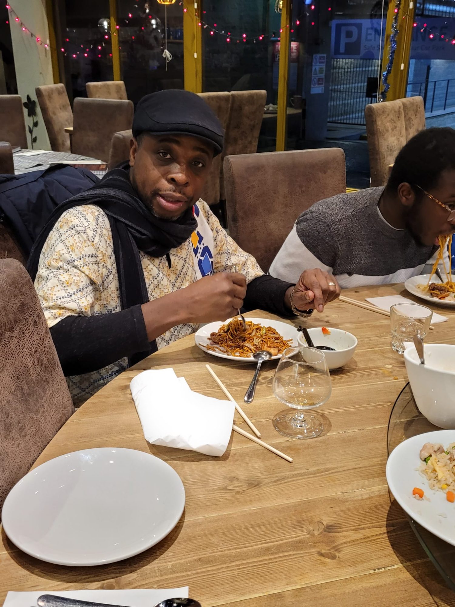 Two men enjoying noodkes at a round table with fairy lights innthe restaurant window