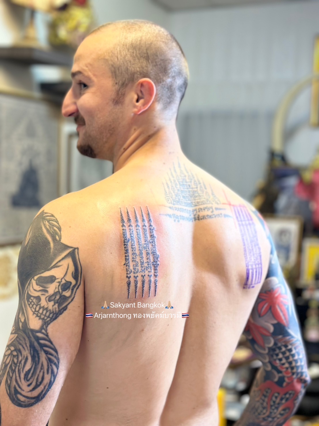 A man with a shaved head is smiling slightly, showing his back and shoulder tattoos. His upper back features fresh Sak Yant tattoos with sacred geometric and script patterns in black ink, and his right arm is covered in colourful sleeve tattoos including a skull design. The background shows a softly blurred temple-like room with framed artwork and golden decorations. The text overlay on the image reads “🙏 Sak yant Bangkok 🙏 Arjanthong ทองพยัคฆ์มาบารมี 🇹🇭”.
