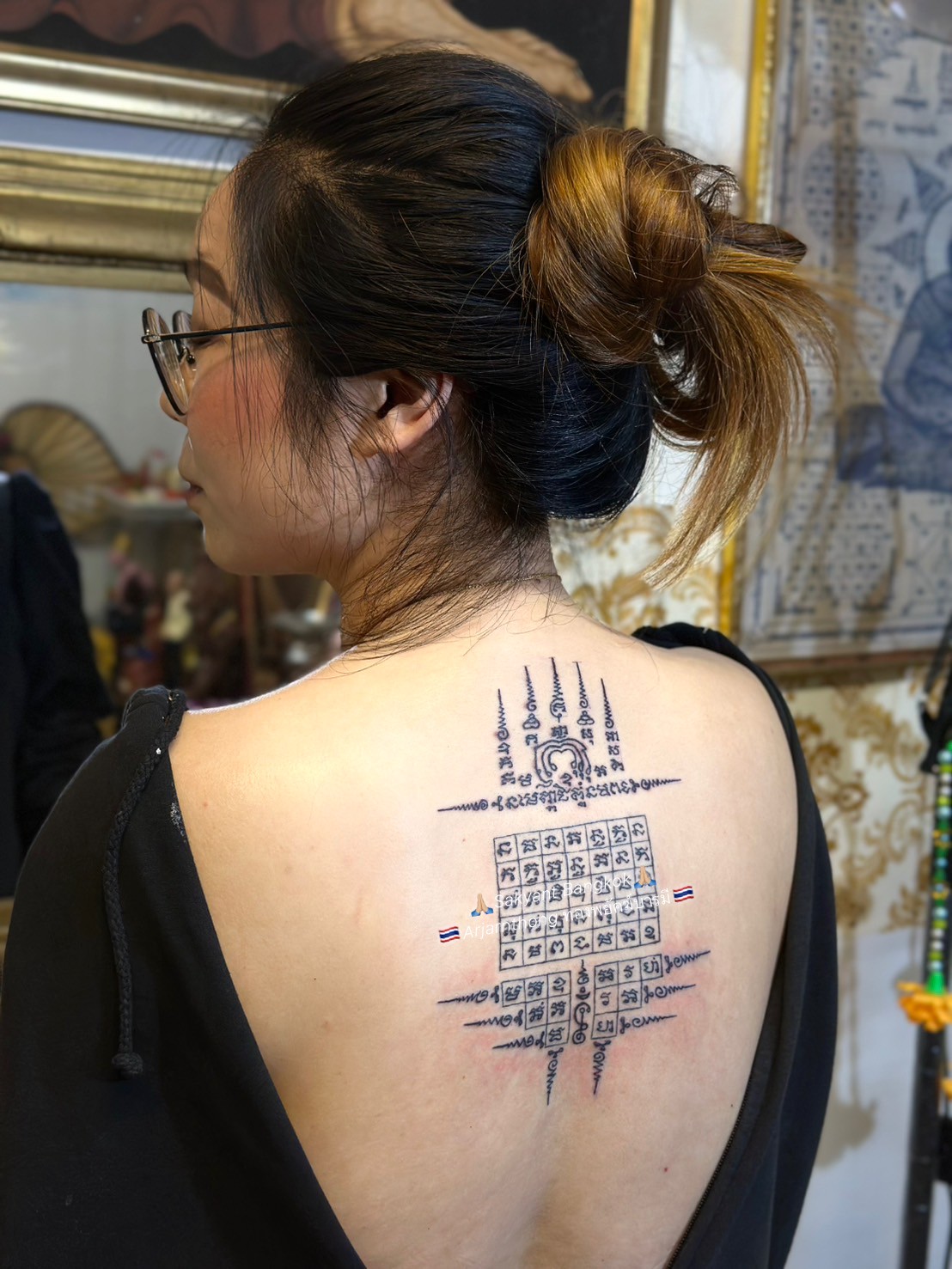A woman with her hair tied in a bun and wearing glasses is seen from behind, revealing a freshly inked Sak Yant tattoo between her shoulder blades. The tattoo is a symmetrical geometric design with intricate script arranged in a square grid and spires pointing in four directions, symbolising protection and spiritual guidance. She is indoors in a ceremonial studio, with framed artwork and gold decorations visible in the background.